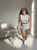 A woman standing in a room with a modern, minimalist aesthetic, wearing a gray one-piece skort with a large cut, paired with a crop top and a belt.