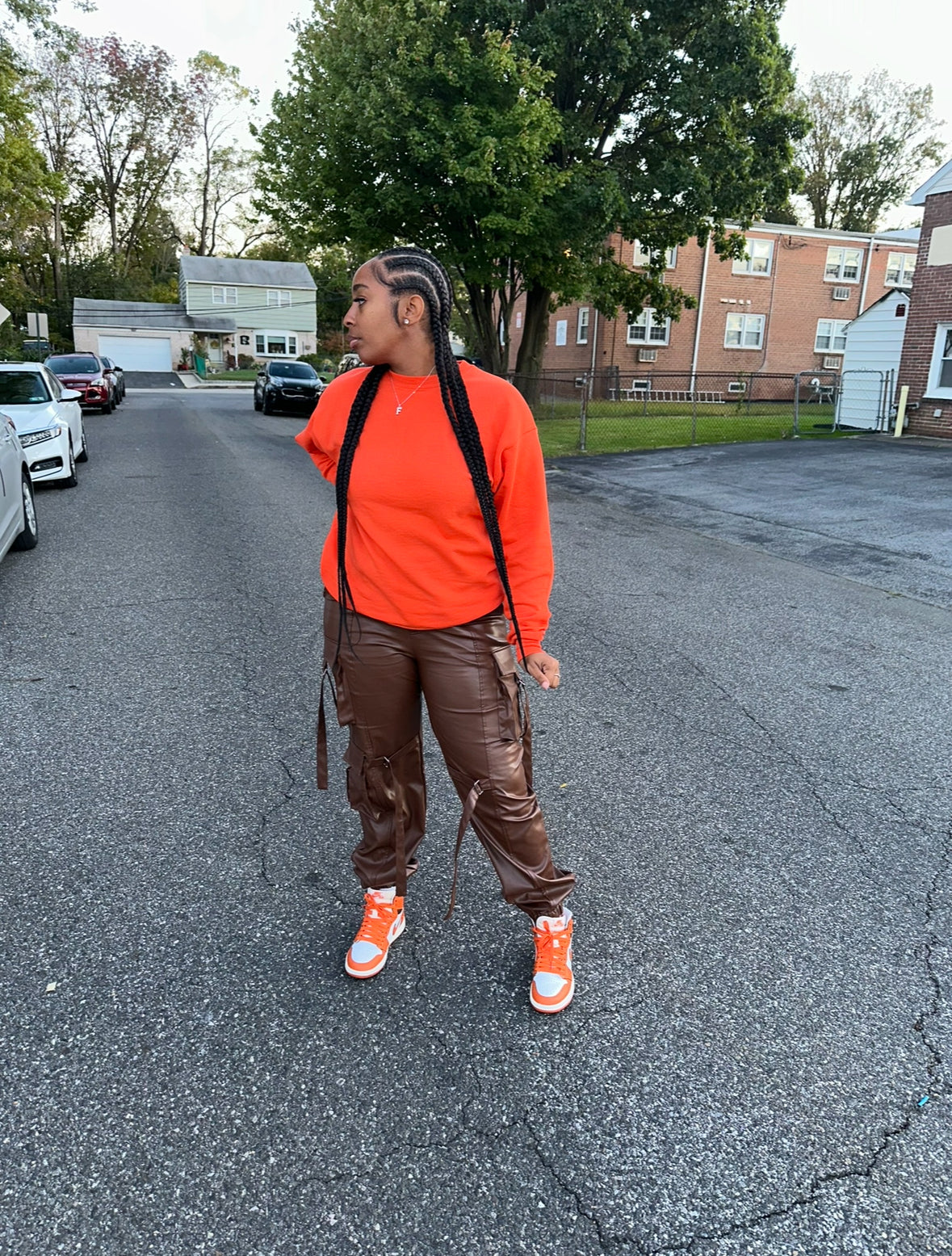 A person wearing chocolate brown faux leather cargo pants with an orange top,  the pants include cargo like pockets, latches, and accessorized placements for a good finish.