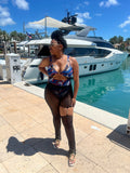 A person wearing mesh-terpiece pants with a multicolored geometric pattern, posing by a marina with boats in the background. The pants are a beautiful cutout, fully see through, and perfect for an addition to the outfit you are wearing.